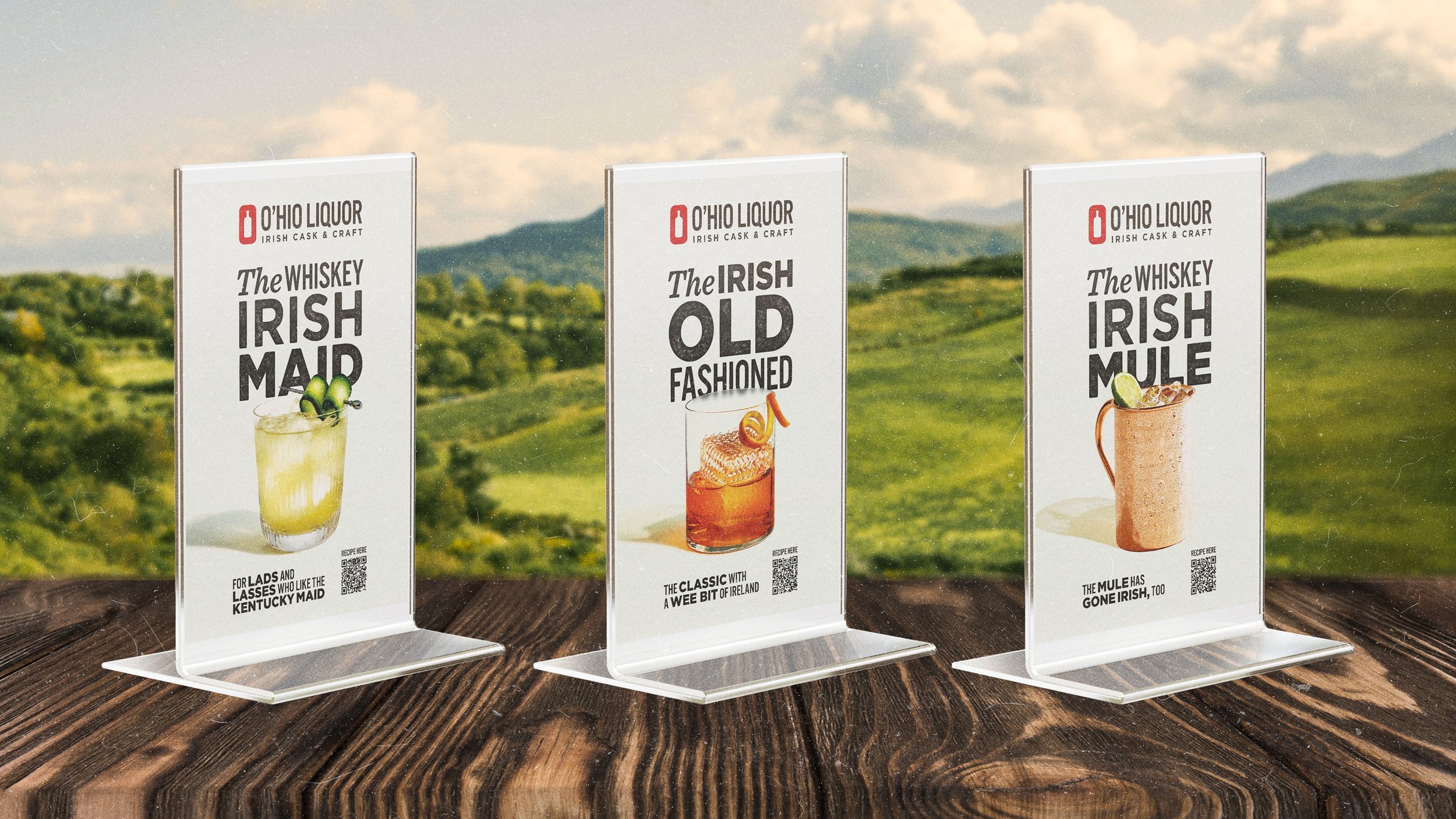 Three vertical advertising signs on a wooden surface, promoting whiskey cocktails with images of drinks and recipes, set against a blurred outdoor landscape with green hills and sky.